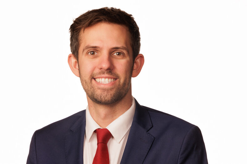 Smiling man with short brown hair in a suit and red tie against a white background.