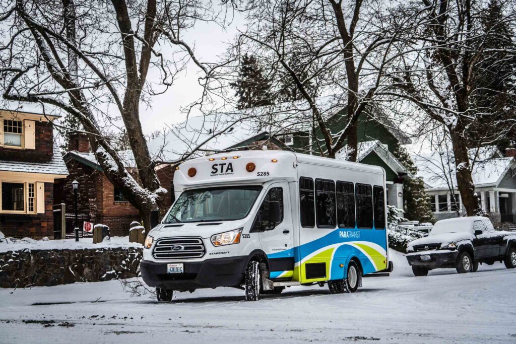 A small bus labeled "STA" drives on a snow-covered residential street lined with trees and parked cars.