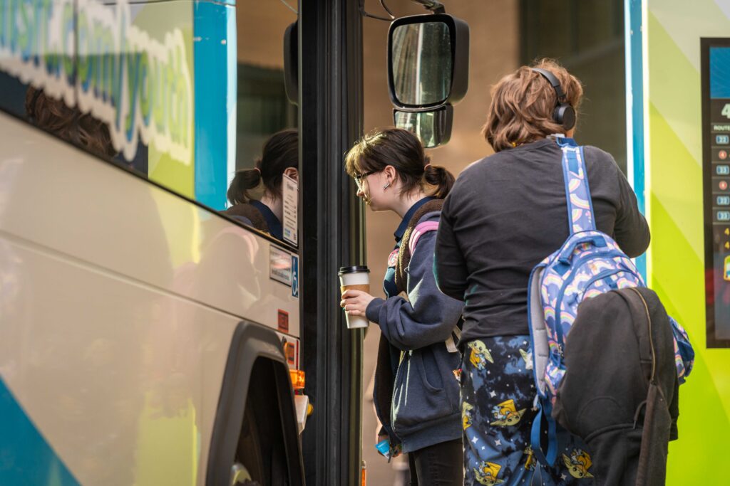 Two people board a bus; one holds a coffee cup, the other has a blue backpack with a yellow pattern.