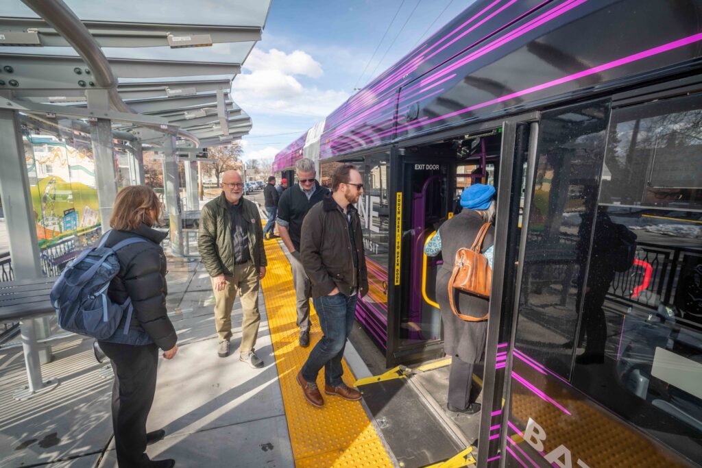 People board a purple-striped city bus at an outdoor transit station.